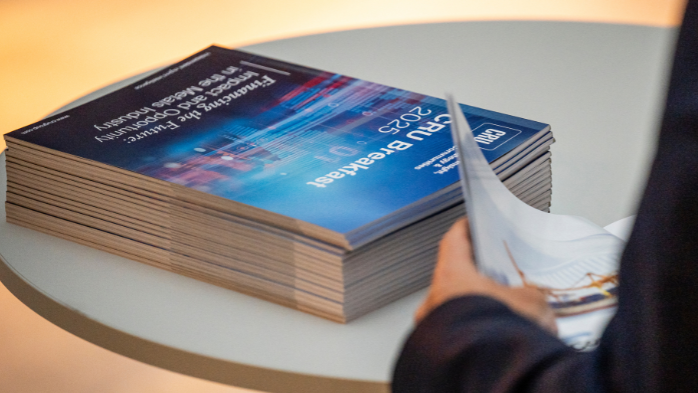 A person standing by a stack of CRU Breakfast Supplement Brochures on a table at the CRU Breakfast event during LME week.